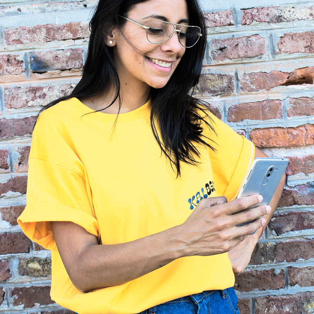 Chica morocha sonriente mientras mira un celular que tiene en la mano. Tiene lentes puestos, remera amarilla de la empresa y jeans