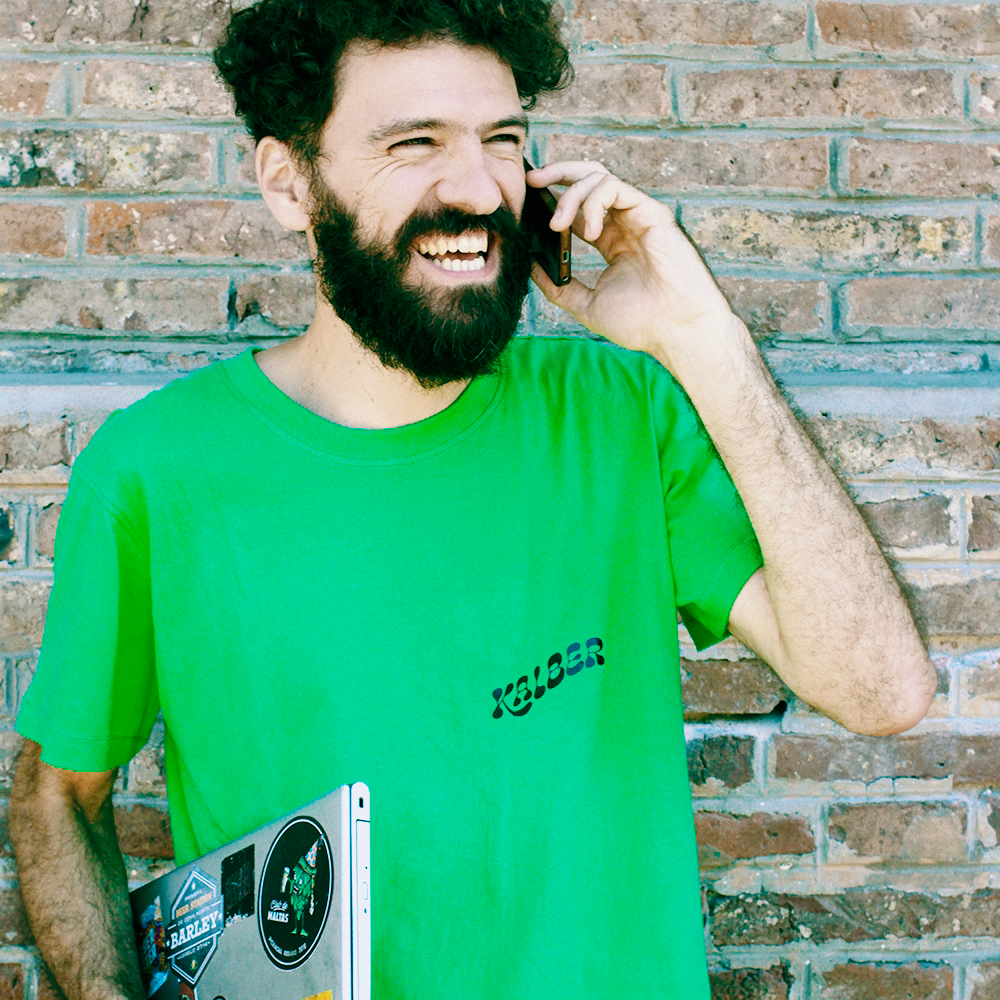 Chico con barba. Remera verde con logo de la empresa. En una mano el celular, en la otra la computadora