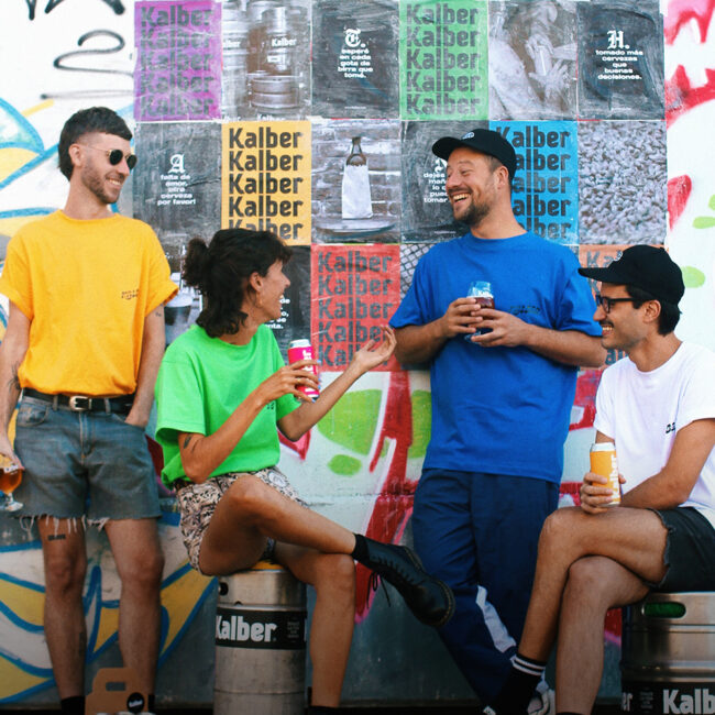 chicos con remers de colres amarillo, verde, azul y blnco, tomando cerveza con pared de fondo con graffitis